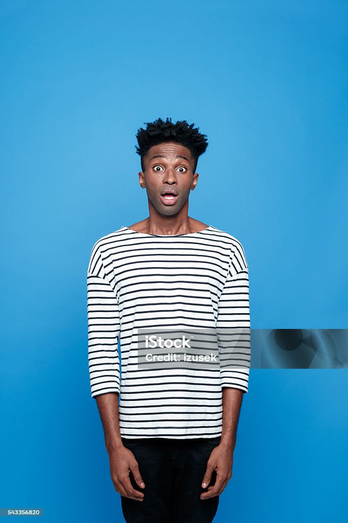 Surprised afro american guy Portrait of surprised afro american young man wearing striped top, staring at camera with mouth open, rolling eyes. Studio portrait, blue background. Adult Stock Photo