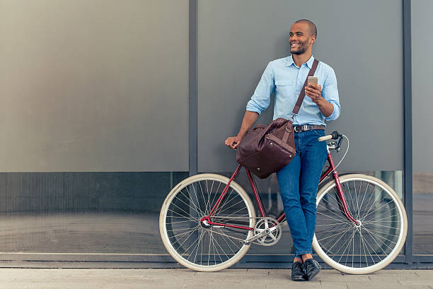 Stylish Afro American businessman Full length portrait of handsome young Afro American man in casual clothes using phone, looking away and smiling while leaning on his bike, standing outdoors urbane stock pictures, royalty-free photos & images