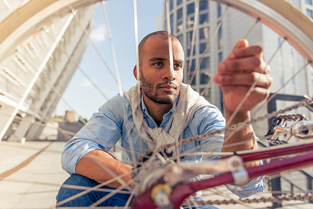 Handsome young Afro American man in casual clothes is examining bicycle spokes, sitting outdoors