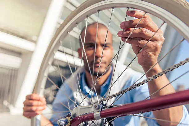Handsome young Afro American man in casual clothes is examining bicycle spokes, sitting outdoors. Focus on hand touching a spoke