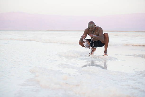 Man on salt beach. Man smeared with healing mud sitting on salt beach of Dead Sea, washing himself with salty water. Body care treatment by natural mineral mud sourced from the salty lake. people covered in mud stock pictures, royalty-free photos & images