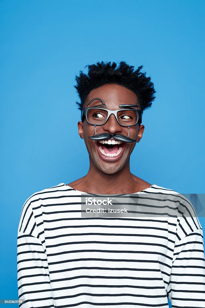 Excited afro american young man wearing funny glasses Portrait of excited afro american guy wearing striped long sleeved t-shirt and funny glasses, laughing. Studio shot, blue background.  Bizarre Stock Photo