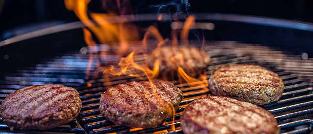 Close-up of juicy burger patties on a ribbed barbecue, flames enveloping the meat.