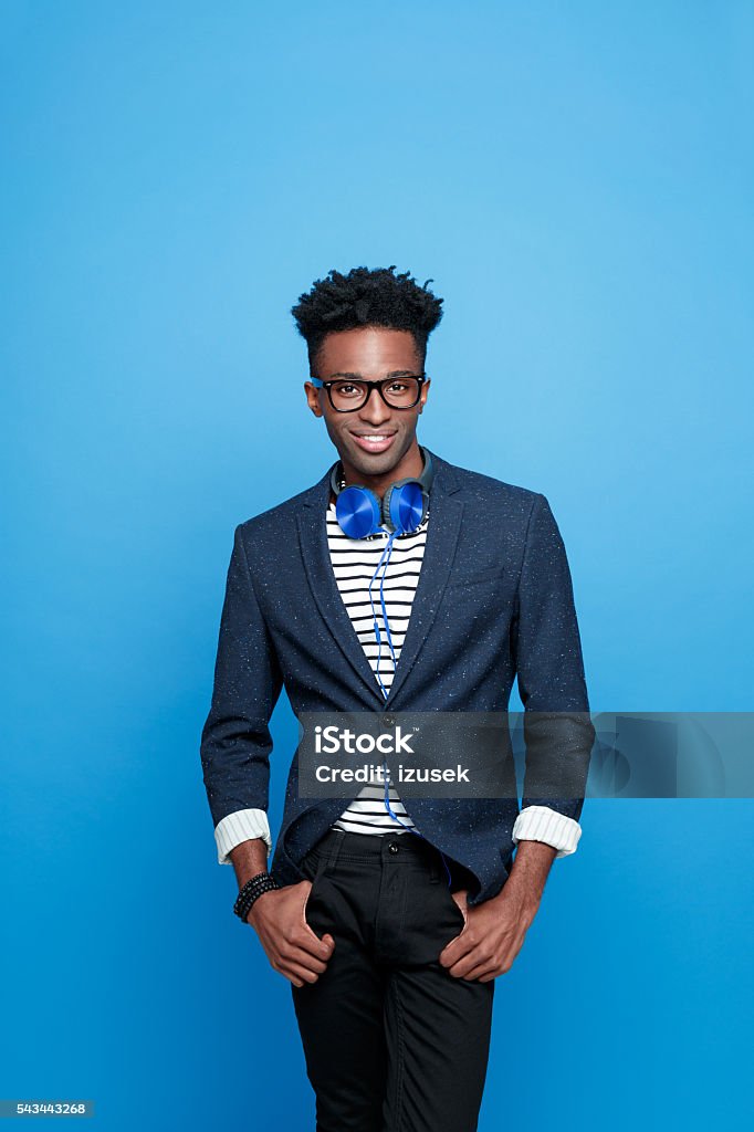 Fashionable afro american guy Studio portrait of fashionable afro american young man wearing striped top, navy blue jacket, nerd glasses and headphone, smiling at camera. Studio portrait, blue background. Colored Background Stock Photo