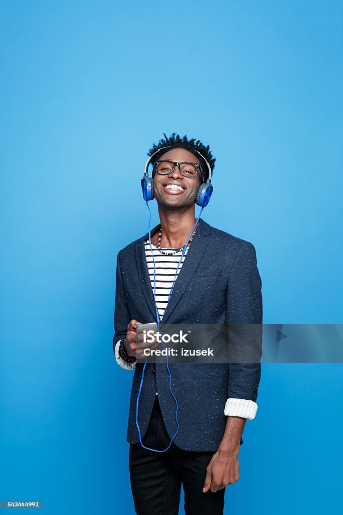 Happy afro american guy using a smart phone Studio portrait of happy afro american young man wearing striped top, navy blue jacket, nerd glasses and headphone, using a smart phone, listening to the music. Studio portrait, blue background. Men Stock Photo
