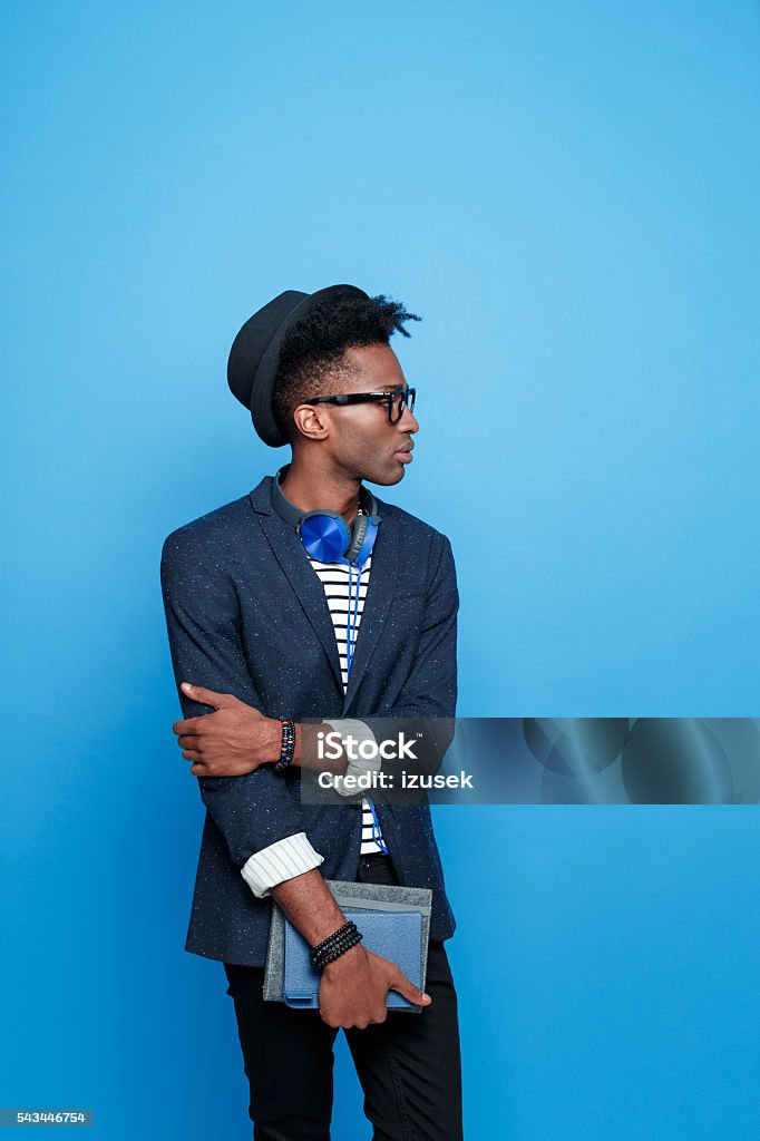 Afro american guy in fashionable outfit, holding notebook Studio portrait of afro american young man wearing striped top, navy blue jacket, hat, nerd glasses and headphone, holding a notebook in hand. Studio portrait, blue background. Adult Stock Photo