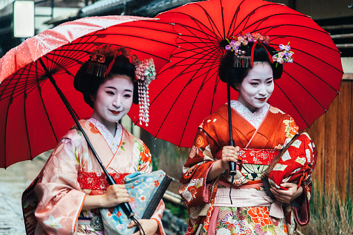 Two maiko geisha walking on a street in Kyoto, Japan during a storm with red umbrella