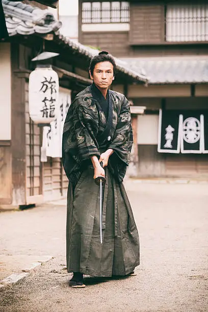 A Japanese Ronin Samurai holds his sword and faces the camera in a defensive posture. Standing on a dirt street in an Edo Period town. Photographed on location in Kyoto, Japan.