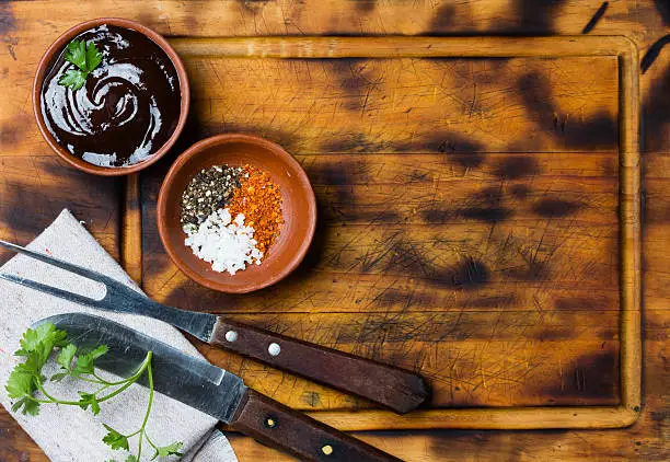 Photo of Barbecue sauce in clay bowl, meat fork, knife, cutting board.