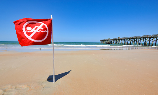 A red no-swimming flag on an empty beach with pier in the background.