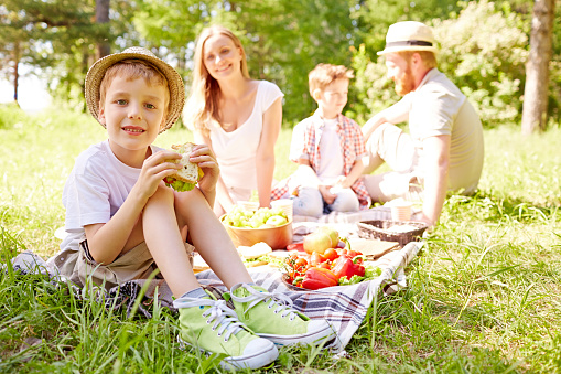 Family at picnic. Child eating sandwich and looking at camera. He and his family spending time in nature. They sitting on picnic blanket. It is perfect for using it in commercial and advertising photography, reports, books, presentation