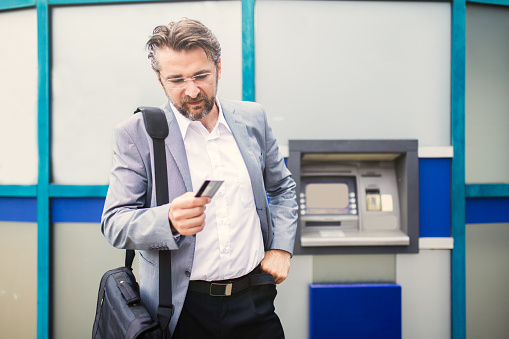 Portrait of a senior businessman standing in front of cash machine. He is holding his credit card worried because it was rejected. Front View.