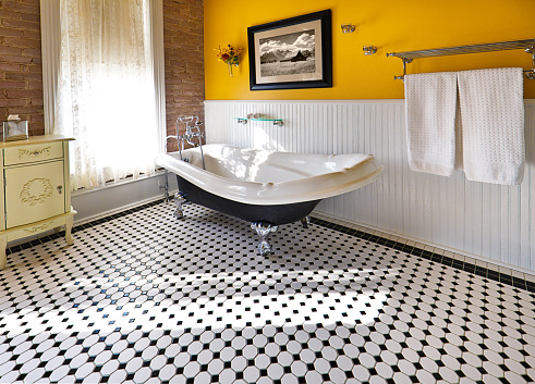 A modern contemporary classic bathroom design, furnished with a classic painted cabinet, sepia toned picture on the wall, exposed brick wall and a window, a claw foot bath tub and black and white tile pattern on the floor. Photographed in horizontal format.