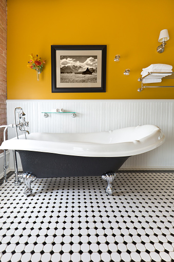 A modern contemporary classic bathroom design, furnished with a classic painted cabinet, sepia toned picture on the wall, exposed brick wall and a window, a claw foot bath tub and black and white tile pattern on the floor. Photographed in vertical format.