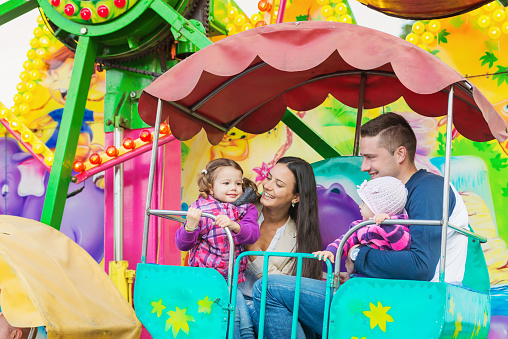 Cute little girls with their mother and father enjoying ride at fun fair, young family, amusement park