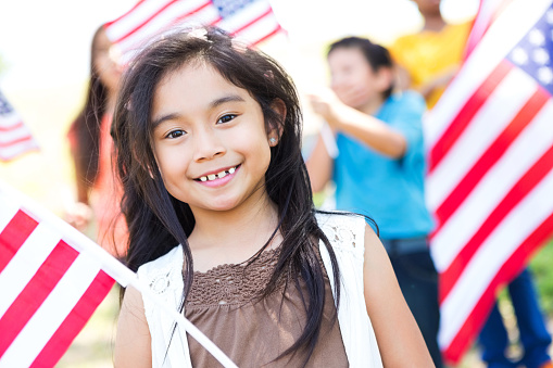 Pretty little girl smiles proudly while holding an American flag. She is attending her neighborhood's 4th of July parade or celebration. Her friends are in the background. She has brown hair and brown eyes.
