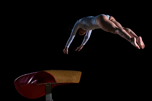 Female gymnast jumping over vaulting horse, black background.