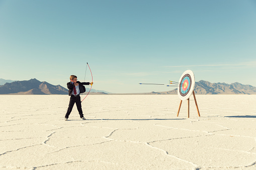 A young boy dressed in business suit, glasses and tie holds shoots a bow and arrow towards a target on the Bonneville Salt Flats in Utah. His business has found success and it mark for the future. He is smiling at the camera in front of blue skies.