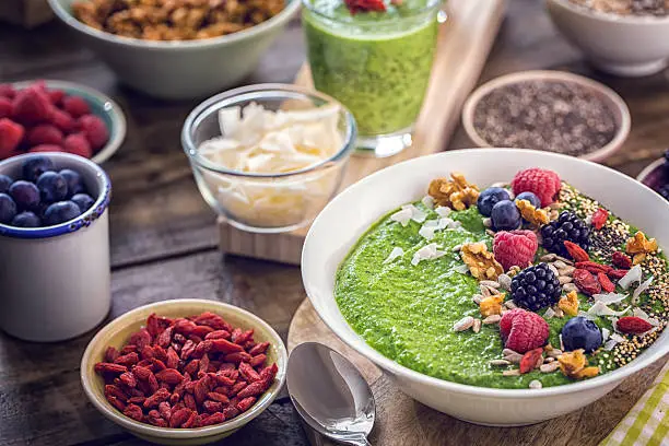 Green breakfast smoothie in bowl with superfoods like chia, quinoa, goji, fresh berries and sunflower seeds.