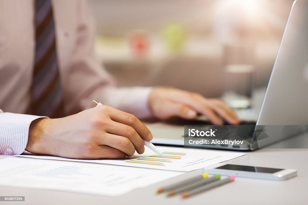 Business Person reading Data on paper Charts and Computer Business Person in Shirt and Tie reading Data Chart on printed Media and working on Laptop Computer with Office Background Telephone and Stationery on Table Analyzing Stock Photo