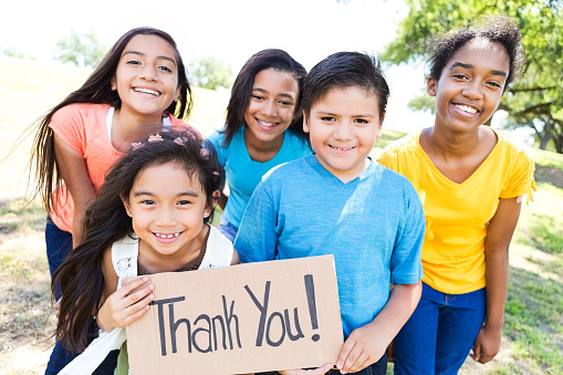 Cute diverse elementary, preteen and teen friends hold 'Thank You!' sign in their neighborhood park.  It is a sunny summer day.