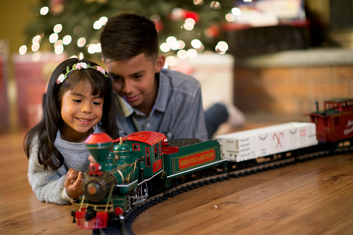 A brother and sister are playing with a toy train on Christmas morning. They are watching the train go around the tracks.