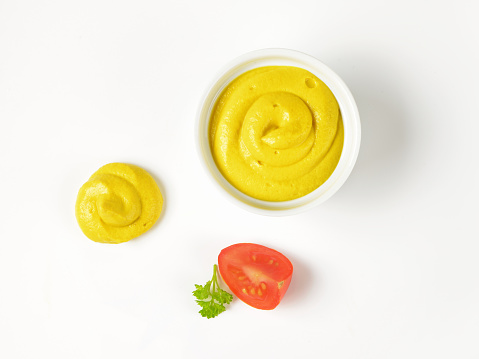 bowl and swirl of yellow mustard and slice of tomato on white background