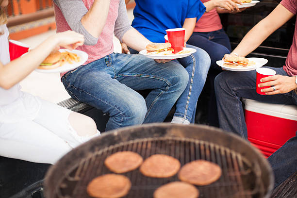 Friends eating burgers at a barbecue Closeup of a group of friends eating hamburgers around a grill during a barbecue tailgate party photos stock pictures, royalty-free photos & images