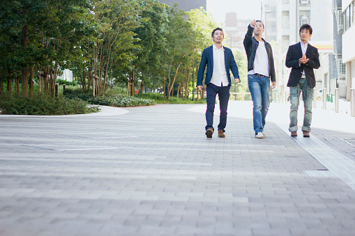 Three Japanese men are walking on a sidewalk in Tokyo. The man of the center is pointing at a forward with his right hand.