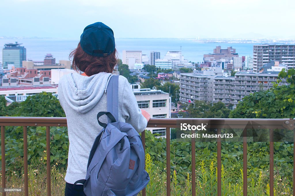 Young woman looking at her town A young woman is standing on slightly higher palce and looking at her town. She is watching architectures and a lake. May be, she is coming back to her home after school. Japanese Ethnicity Stock Photo