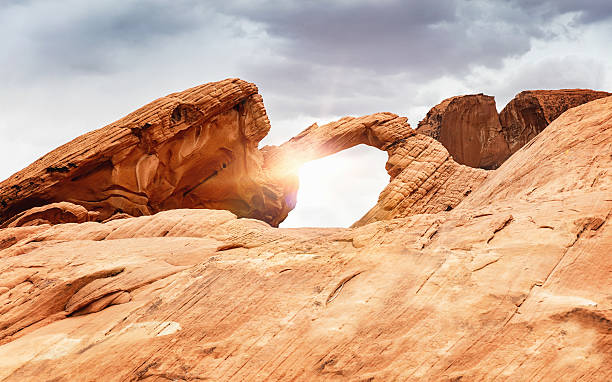 Arch Rock against the Sun. Sunbeam shining though crimson colored natural arch rock sandstone formations at the famous Valley of Fires State Park, The Valley of Fire derives its name from red sandstone formations, formed from great shifting sand dunes during the age of dinosaurs.Valley of Fire State Park, Nevada, USA.