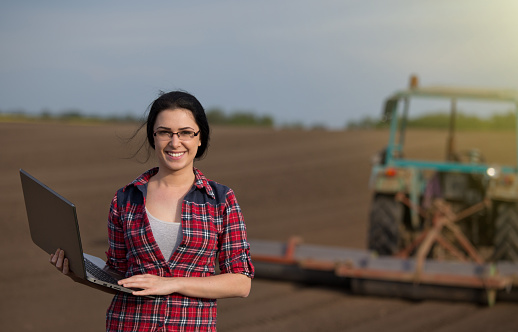 Young happy farmer girl standing on field with laptop in spring time and looking at camera. Tractor working in background