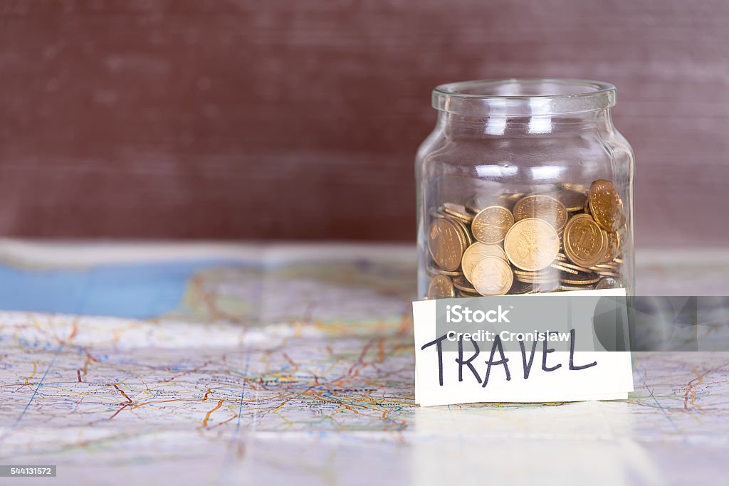 Coins in a jar with travel sign on a map. Coins in a jar with travel sign on a map against vintage brown wooden background. Copy space on the left. Concept of travelling. Budget Stock Photo