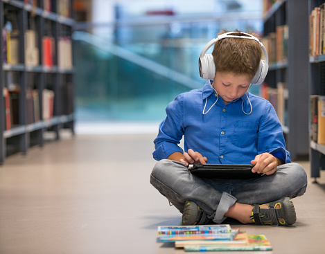 Schoolboy with headphones and tablet computer sitting on library floor.