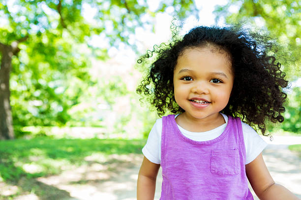 Beautiful little girl in the park Cute African American little girl walks in the park on a sunny day. The wind is blowing her curly black hair. She has brown eyes and is wearing a purple tank top with a white t-shirt underneath. Grass and trees are blurred in the background. Copy space available. cute black babys stock pictures, royalty-free photos & images