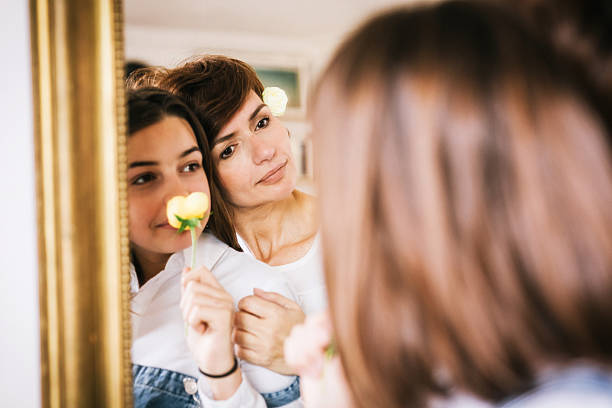 Mother and Teenage girl daughter with flower posing arm in arm in front of a mirror smiling happily at their reflection
