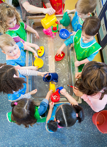 Overhead view of a class of nursery children playing at a water table. They are all wearing aprons and using plastic toys.