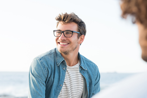 Handsome man smiling at the beach and looking away. Portrait of a cheerful guy enjoying summer vacation. Handsome man with eyeglasses in casual relaxing outdoor with friend.
