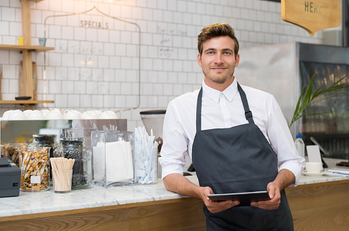 Successful small business owner holding digital tablet and looking at camera. Happy smiling waiter with apron and digital tablet leaning on counter. Portrait of young entrepreneur of coffee shop posing.