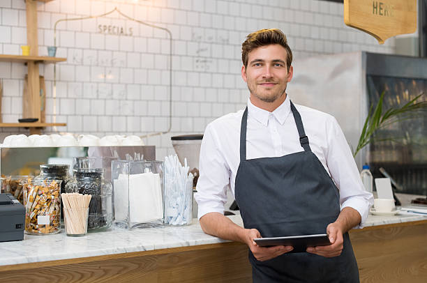 Small business Successful small business owner holding digital tablet and looking at camera. Happy smiling waiter with apron and digital tablet leaning on counter. Portrait of young entrepreneur of coffee shop posing. waiter stock pictures, royalty-free photos & images