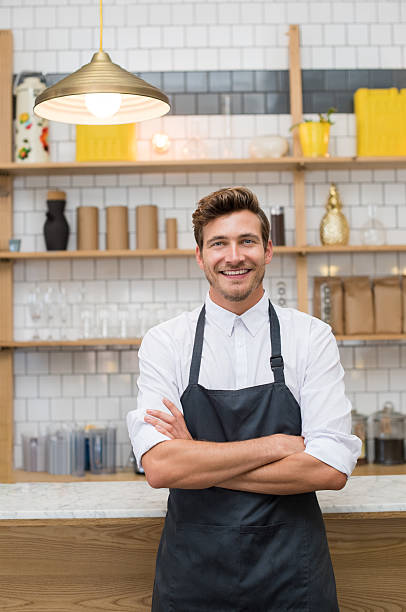 Happy young entrepreneur Portrait of a smiling young waiter leaning at counter of coffee shop with arms crossed. Successful owner with apron looking at camera. Young cook standing in kitchen. Small business concept. man cooking apron stock pictures, royalty-free photos & images
