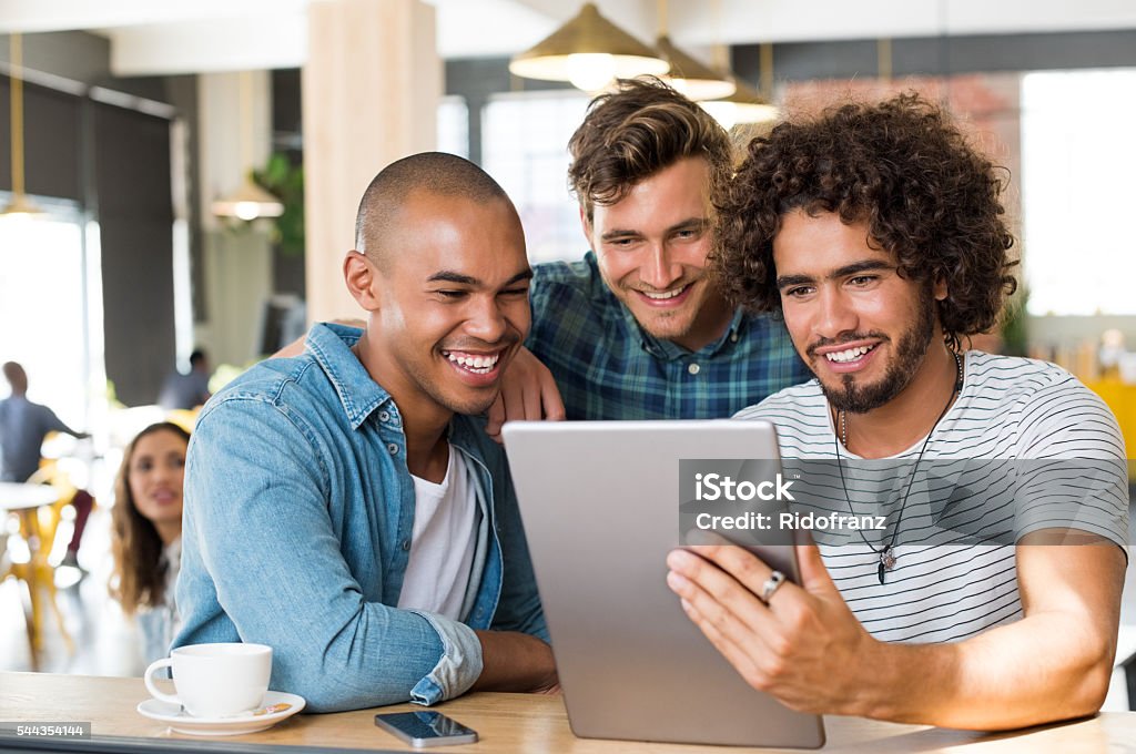 Friends using tablet Young guys in casual clothes smiling and discussing ideas using tablet at cafe. Group of friends looking at digital tablet. Happy multiethnic young men watching video on digital tablet in a wifi cafe. Friendship Stock Photo