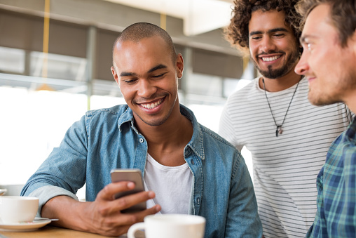 Cheerful african man using smartphone at coffee shop with his friends. Young man with friends reading a message on smartphone. Smiling guy texting a phone message.