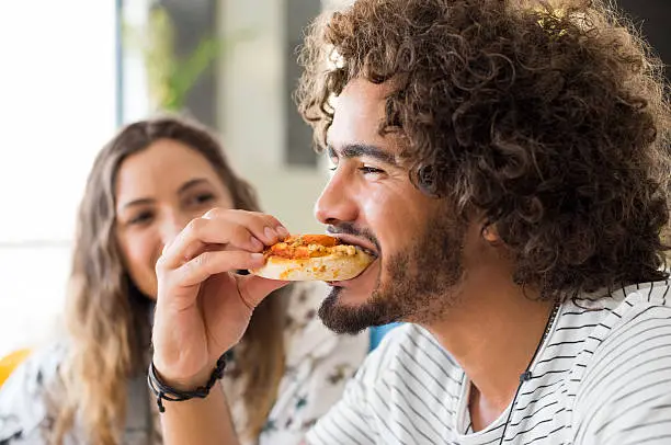 Close up face of a young african man eating a pizza in a coffee shop. Happy guy with friends enjoying brunch in a cafeteria. Portrait of a multiethnic young man biting pizza.