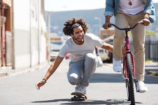 Closeup of a happy young man skateboarding with help of friend bicycle. Smiling guys having fun with skate board and cycle. African man riding a bike while his friend attacking with skateboard.