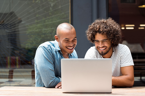Two happy friends using laptop outdoor. Happy african young men laughing while watching laptop screen. Two young smiling guys using a laptop in a cafe bar.