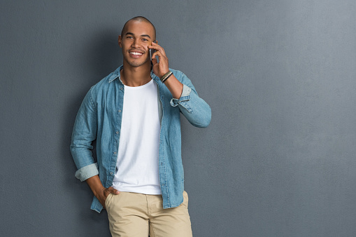 Young handsome man talking over phone and looking at camera. Young man using smartphone while leaning against grey wall. Portrait of happy casual african guy talking on the phone on grey background with copy space.