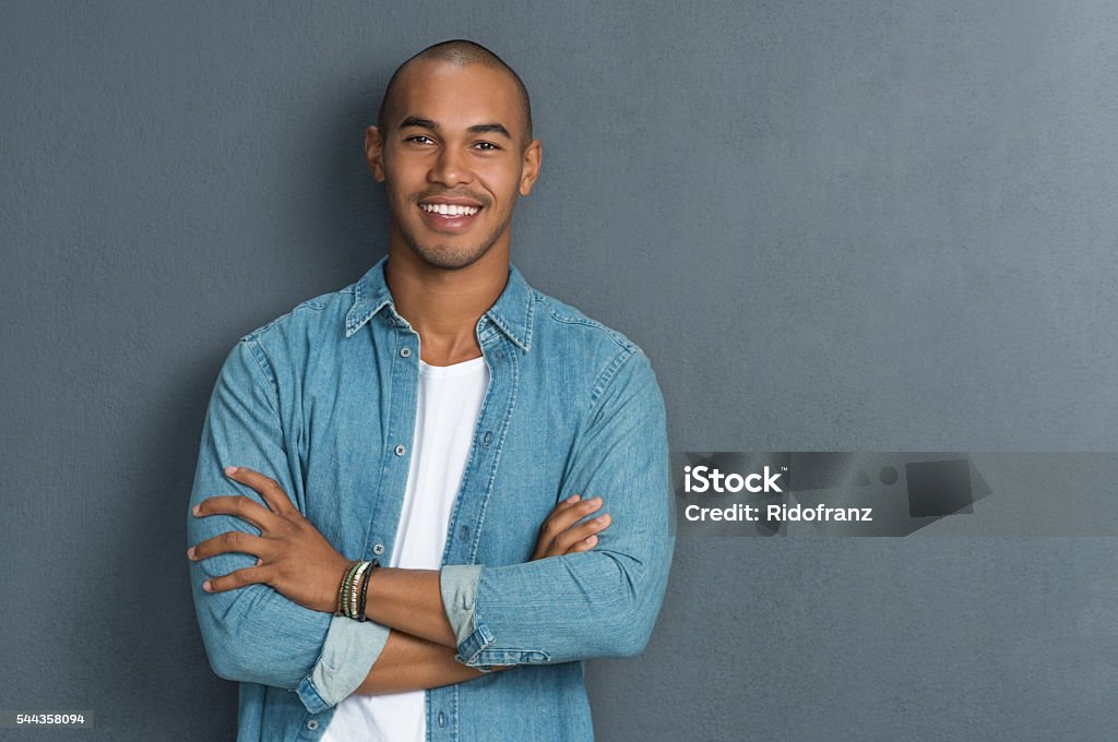 Cool african man Portrait of handsome man standing with arms crossed in smart casual clothing against a grey wall. Smiling young african guy looking at camera leaning on grey background with copy space. Portrait of a satisfied man looking at camera. Men Stock Photo