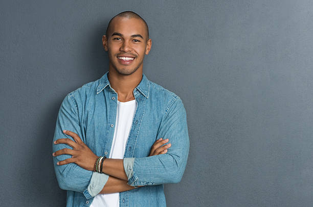 Cool african man Portrait of handsome man standing with arms crossed in smart casual clothing against a grey wall. Smiling young african guy looking at camera leaning on grey background with copy space. Portrait of a satisfied man looking at camera. handsome people stock pictures, royalty-free photos & images
