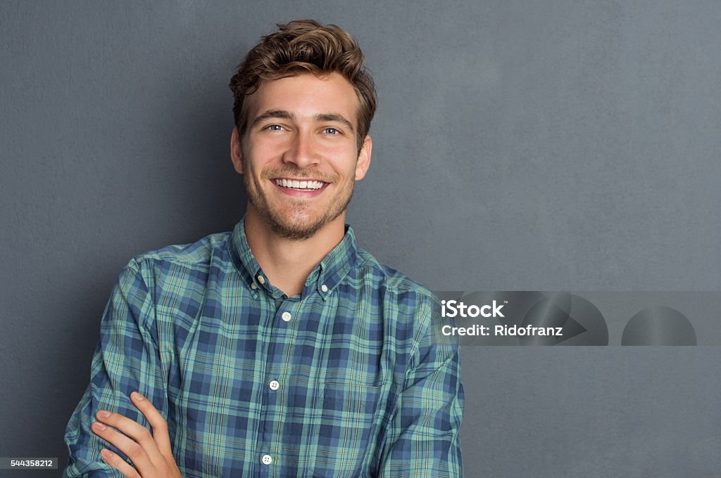 Happy laughing man Young handsome man leaning against grey wall with arms crossed. Cheerful man laughing and looking at camera with a big grin. Portrait of a happy young man standing with crossed arms over grey background. Men Stock Photo